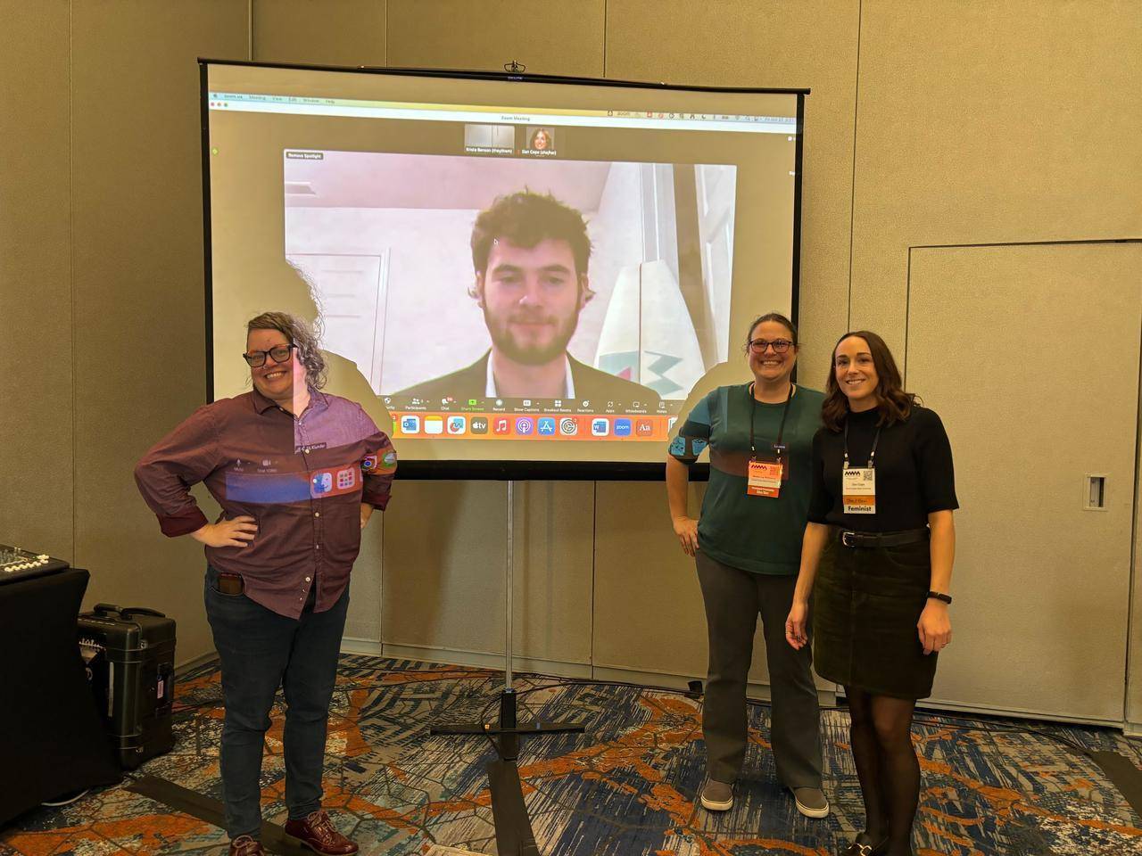 Photo of two women standing in front of a screen showing a male student on zoom for a presentation
